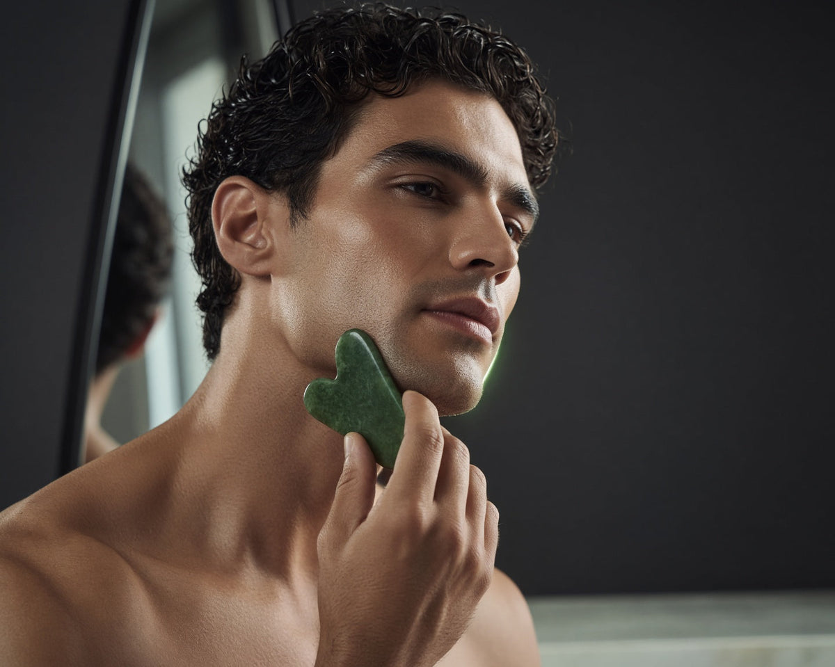 Man holding a jade gua sha stone to his jawline in front of a mirror, part of the GLOWRYZE 5-minute daily facial ritual