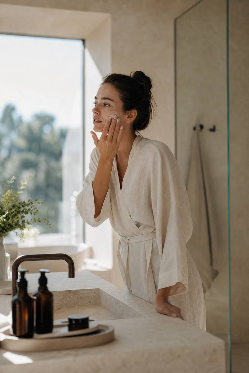 Woman applying facial cream in front of a sunlit bathroom mirror